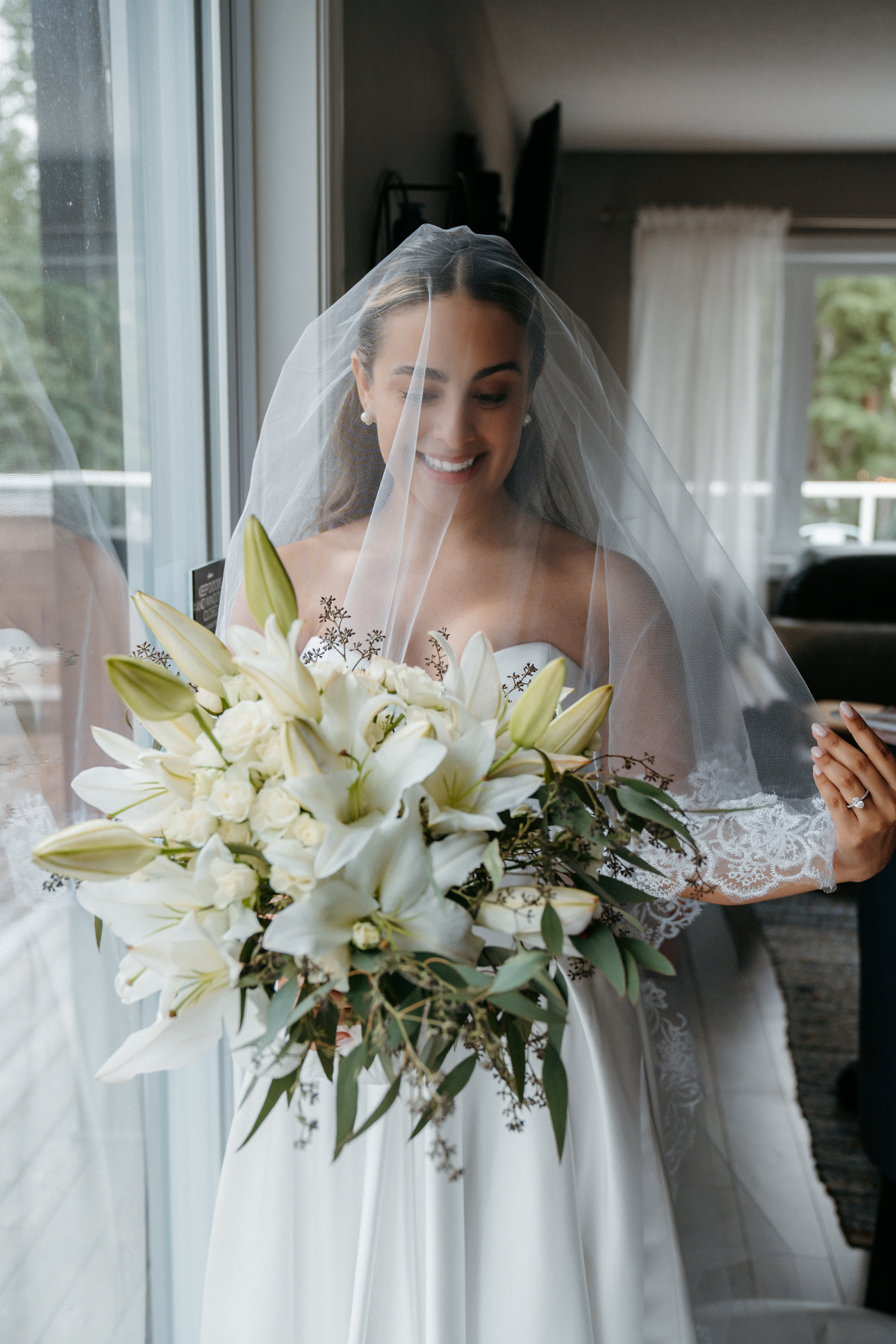 Bride smiling under her veil holding a lush white lily and rose bouquet by Borealis Blooms